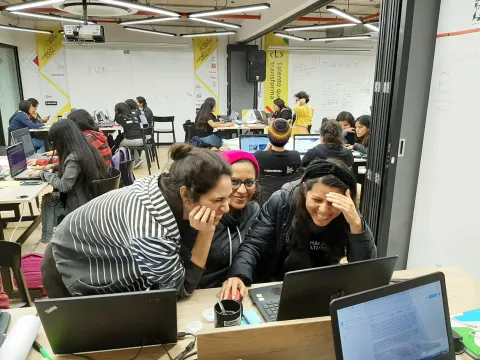 Classmates working and laughing at their laptops during the hackathon, with the event banners behind them.