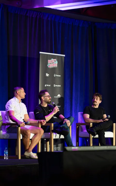 Three speakers seated on stage during a panel, under blue and purple light, with the React Miami banner behind them.