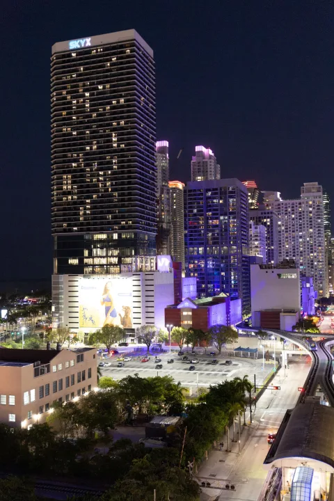 Miami towers lit up at night, seen from above with a metro track curving below.