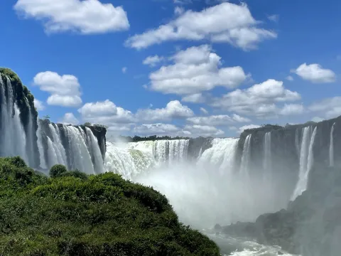 Wide multi-tier waterfalls throwing spray, with green vegetation in the foreground and scattered cloud in a blue sky.