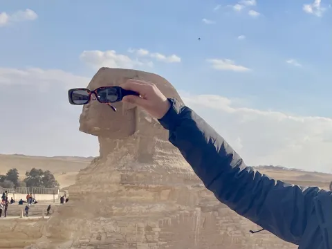 A hand holding a pair of sunglasses up in front of the Great Sphinx, as if putting them on it.