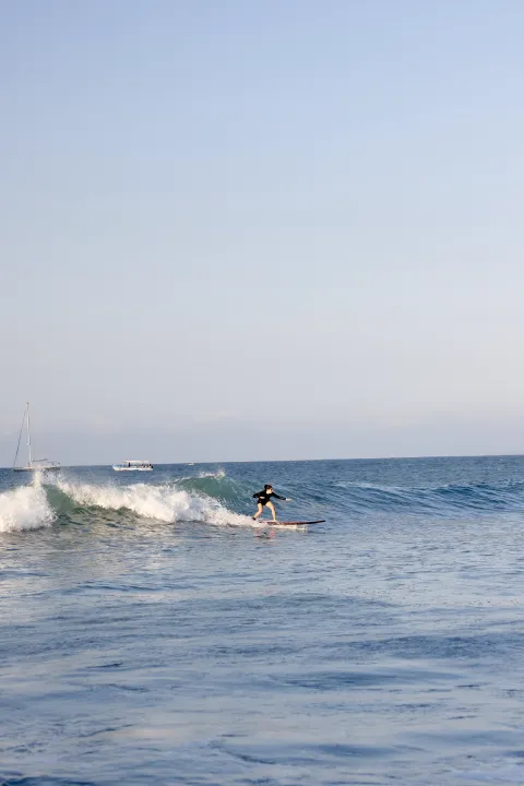 A lone surfer riding a small breaking wave on a wide calm sea, with two boats far off on the horizon.