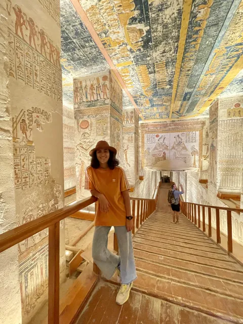 Gabriela standing on a wooden walkway inside a painted tomb corridor, hieroglyphs and figures covering the walls and ceiling.